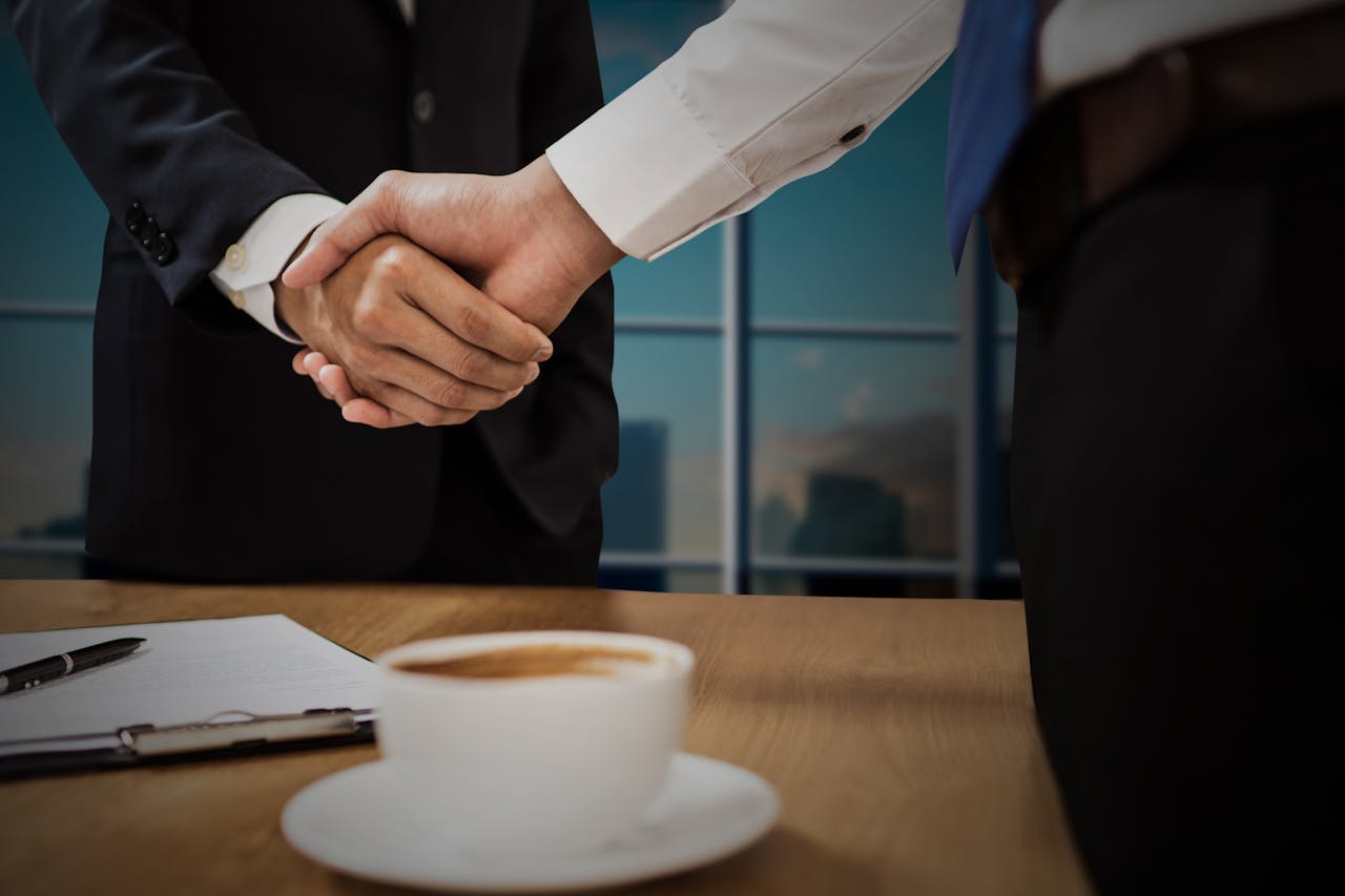 Two professionals shaking hands across a desk with cityscape in the background.